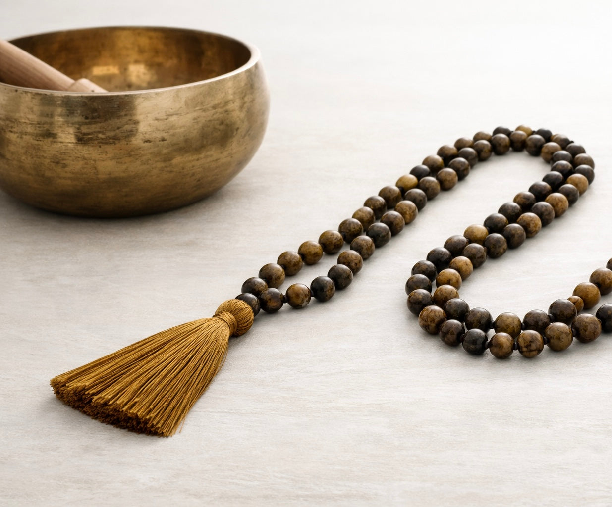 Mala sautoir avec perles naturelles d'oeil de tigre et pompon coton marron posé sur un tissu ivoire, à côté d'un bol tibétain en bronze