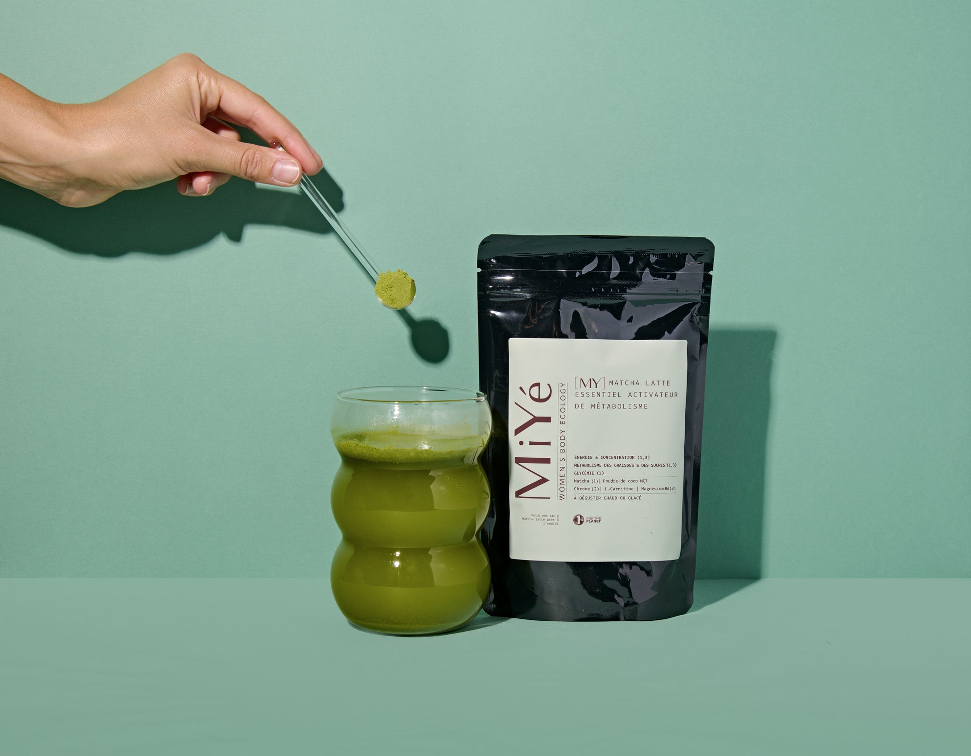 Mains tenant une cuillère doseuse avec poudre de thé vert matcha, au-dessus d'un verre rempli de matcha latte, à côté d'un sachet de My Matcha Latte MiYé
