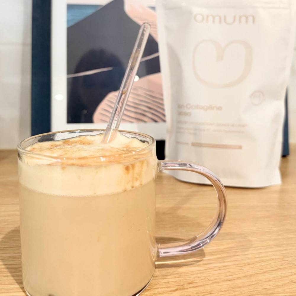 Tasse et paille en verre contenant le complément alimentaire Mon collagène cacao d'Omum, avec son sachet, posés sur une table en bois clair