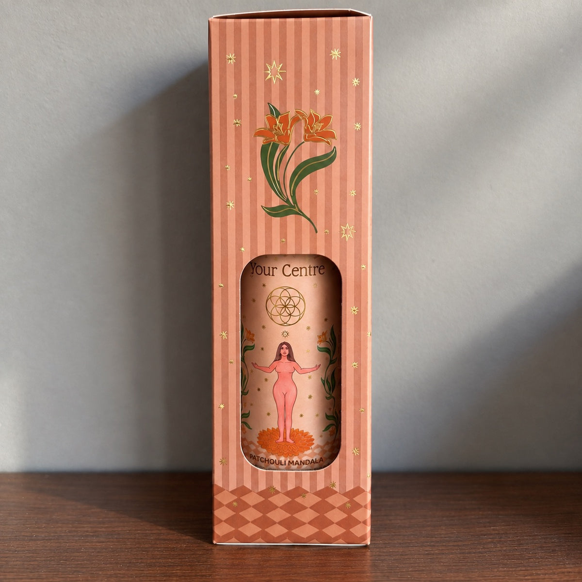 Étui rayé terracota du spray d'ambiance Find Your Centre Carnatia, orné de fleurs dorées et d'étoiles, avec fenêtre découpée laissant apparaître le flacon patchouli mandala et son illustration de femme les bras ouverts.