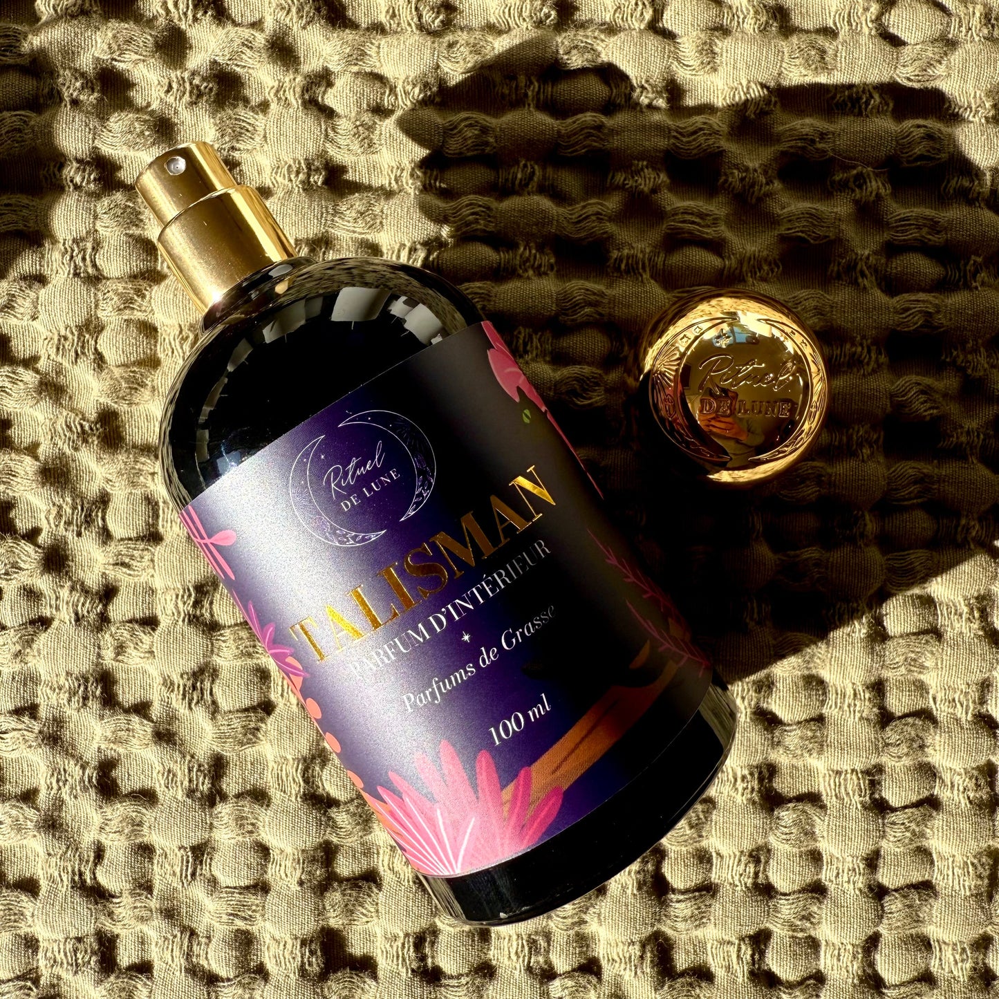 Flacon pompe parfum d'intérieur Talisman  Rituel de lune, parfums de grasse 100ml, en verre brun et étiquette violette, or et rose, avec son bouchon doré posé à côté sur un linge en coton kaki