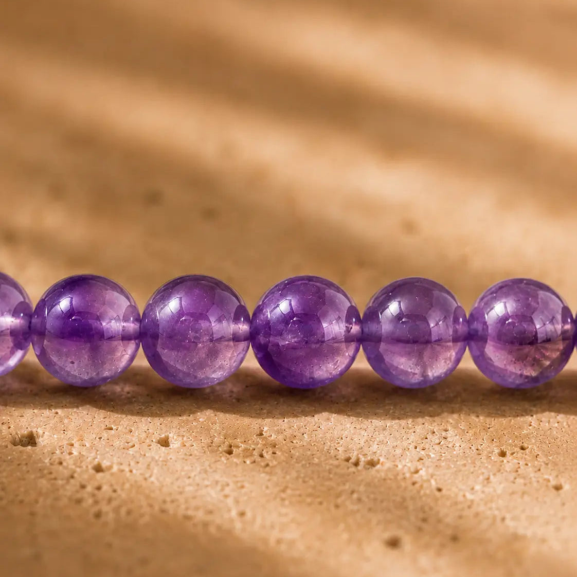 Gros plan sur les perles rondes d’améthyste violettes et translucides du bracelet Medium Jolis Baume sur une pierre beige

