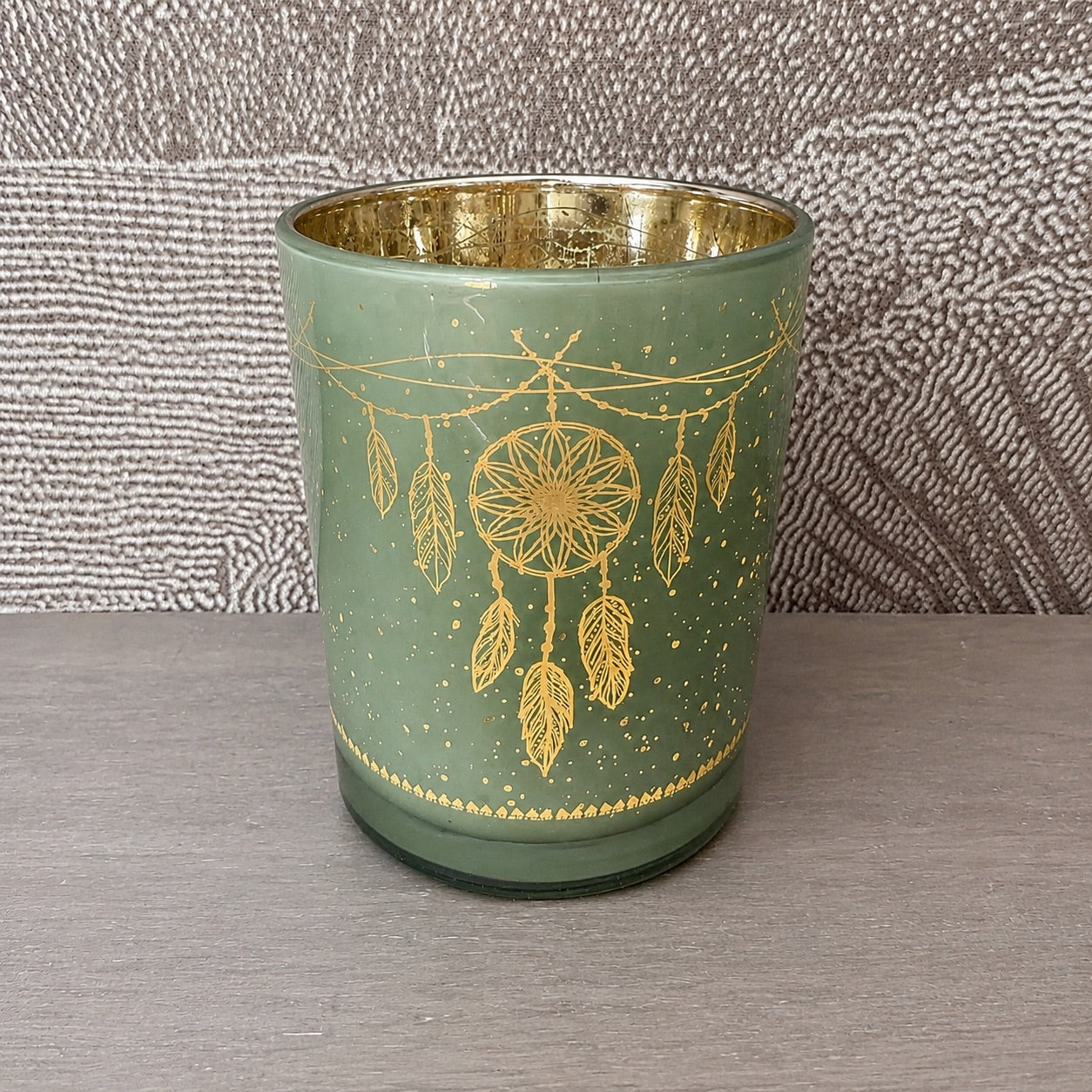 Photophore attrape-rêves en verre vert sauge posé sur une étagère en bois gris, motif attrape-rêves doré avec plumes, intérieur doré métallisé visible, décoration énergétique
