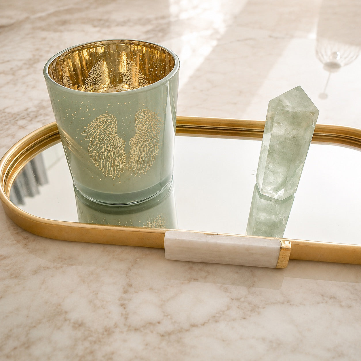 Photophore cylindrique métallisé en verre vert sauge avec motif ailes d'ange dorées et étoiles posé sur un plateau miroir et doré, accompagné d'un cristal vert en pointe