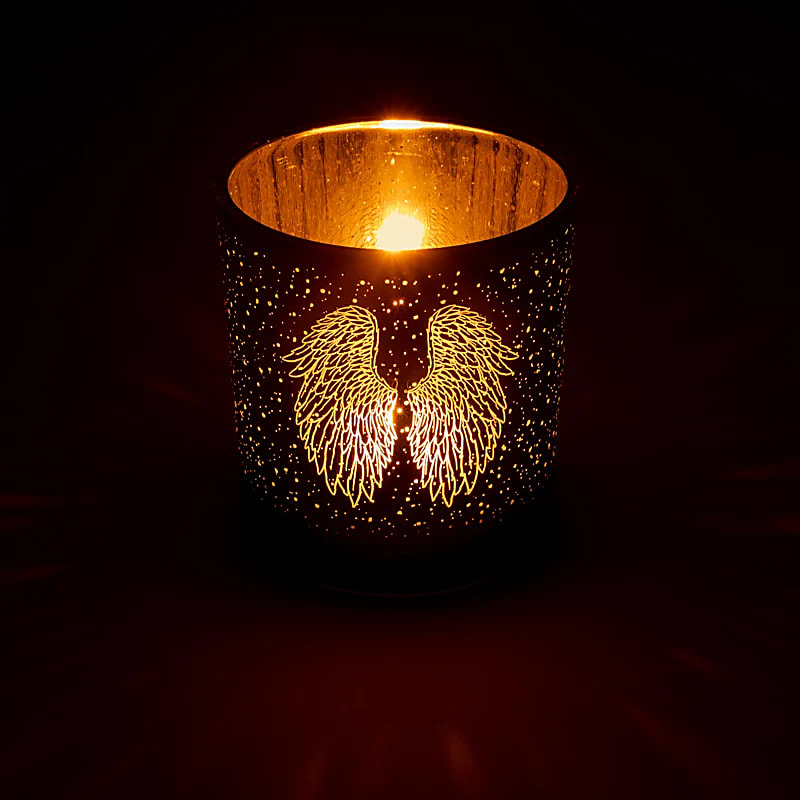 Photophore cylindrique métallisé avec motif ailes d'ange dorées, illuminé par une bougie, projetant une lumière chaude et dorée parsemée d'étoiles