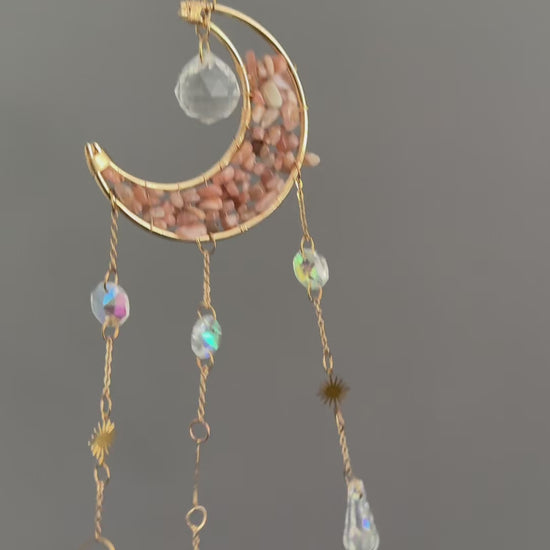 Vidéo de présentation d'un attrape-soleil doré en forme de croissant de lune, garni d'éclats de pierre de lune rose, avec trois chaînes pendantes ornées de soleils dorés et de pendentifs en cristal de roche facetté