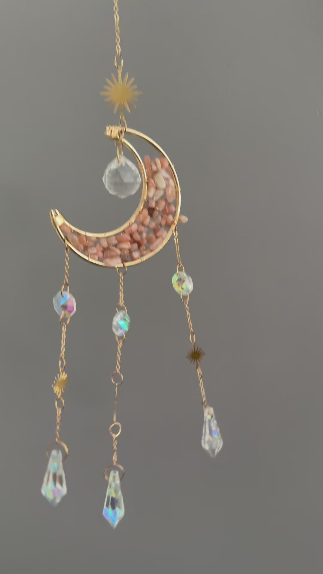 Vidéo de présentation d'un attrape-soleil doré en forme de croissant de lune, garni d'éclats de pierre de lune rose, avec trois chaînes pendantes ornées de soleils dorés et de pendentifs en cristal de roche facetté