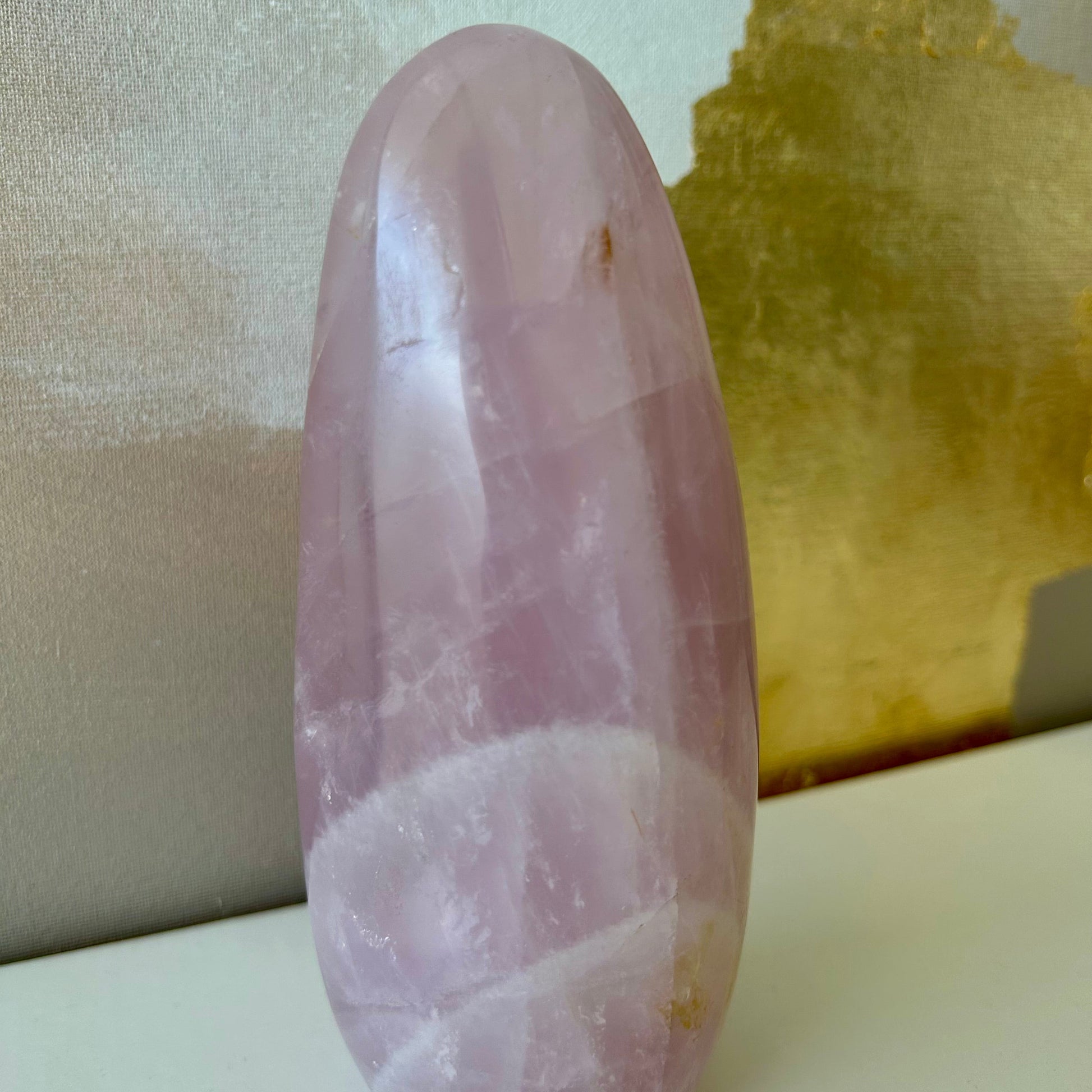 Pierre naturelle de Quartz rose en forme de dolmen posée sur étagère blanche et toile dorée et beige en arrière-plan