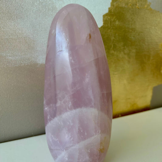 Pierre naturelle de Quartz rose en forme de dolmen posée sur étagère blanche et toile dorée et beige en arrière-plan