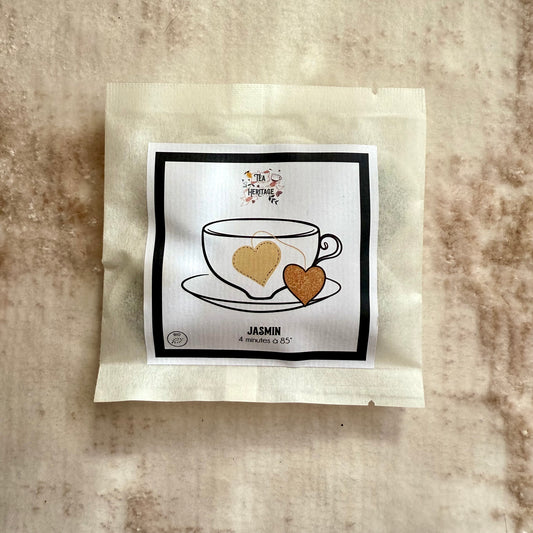 Pochette de thé au jasmin avec une étiquette avec la marque Tea Heritage, une tasse de thé dessinée et deux coeurs or reliés par un fil, sur fond beige