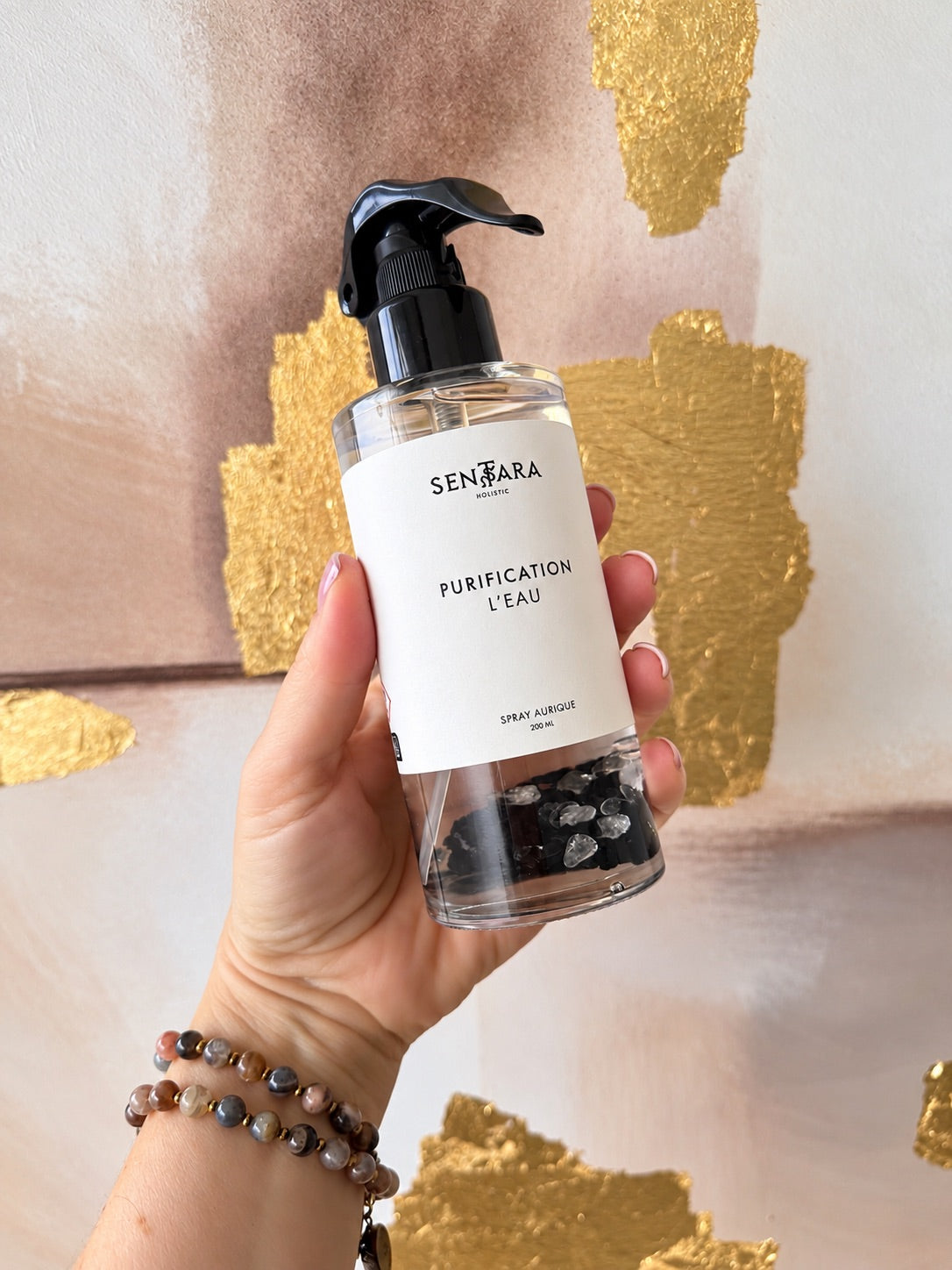 Une main portant des bracelets de pierre de lune tient le spray aurique Purification L’Eau 200 ml de Sentara Holistic devant un fond aux touches dorées