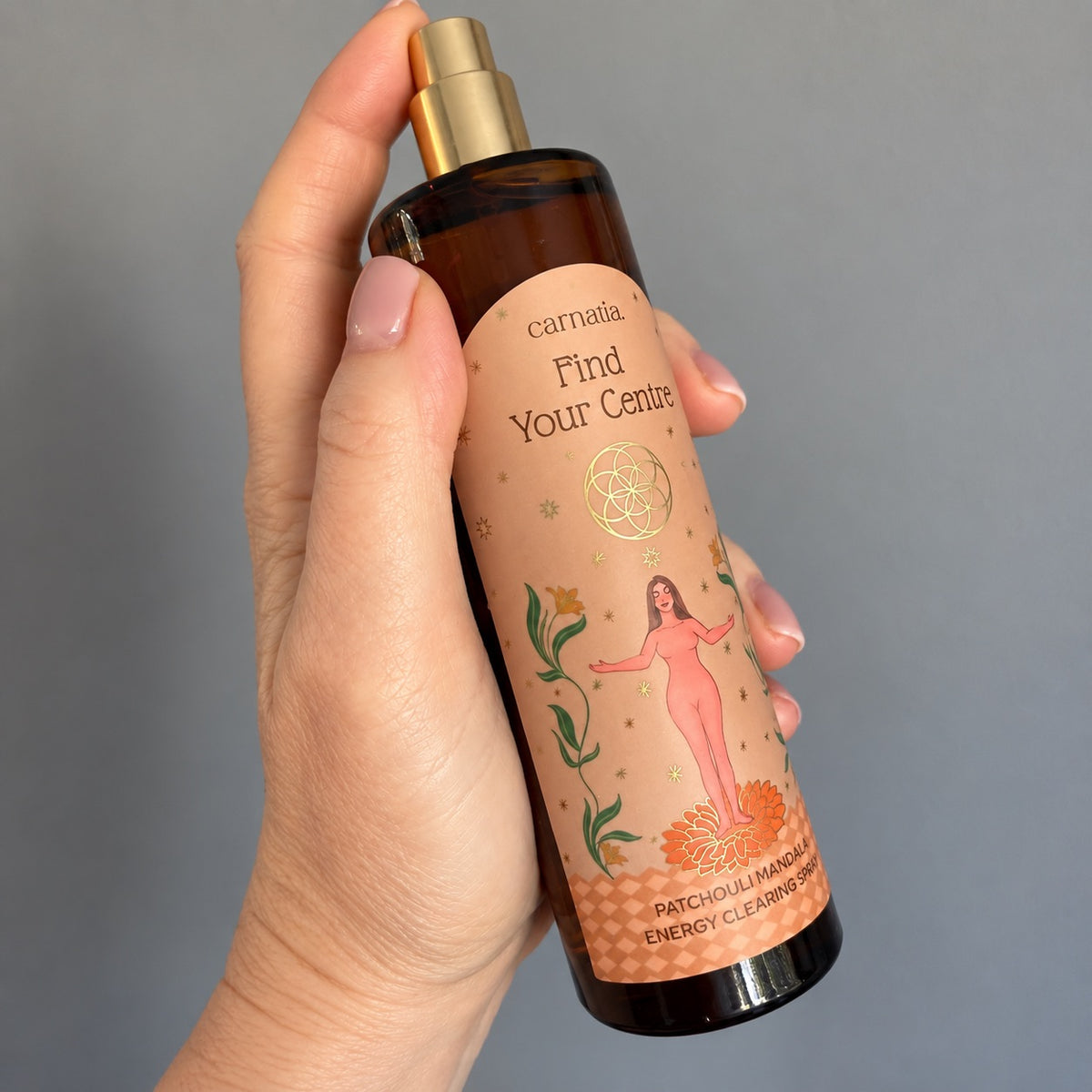 Main tenant le flacon vaporisateur ambré Carnatia Find Your Centre, étiquette rose ornée d'une fleur de vie dorée et d'une illustration de femme les bras ouverts, spray d'ambiance énergétique patchouli mandala.
