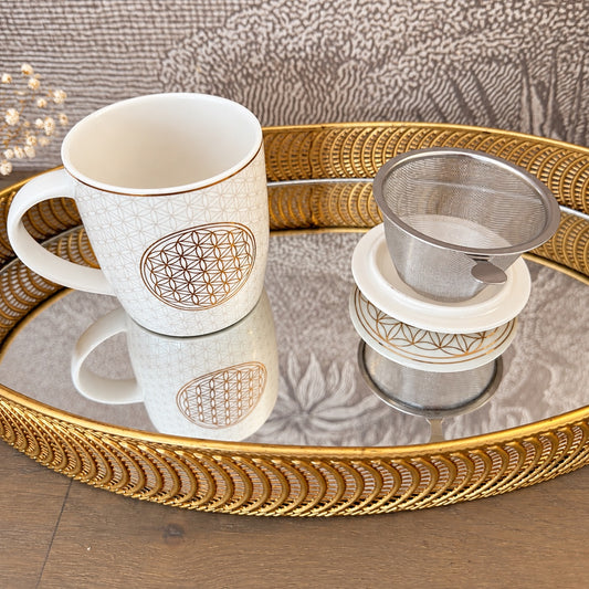 Tasse blanche ornée d’une Fleur de Vie dorée avec son infuseur posé sur un plateau miroir doré