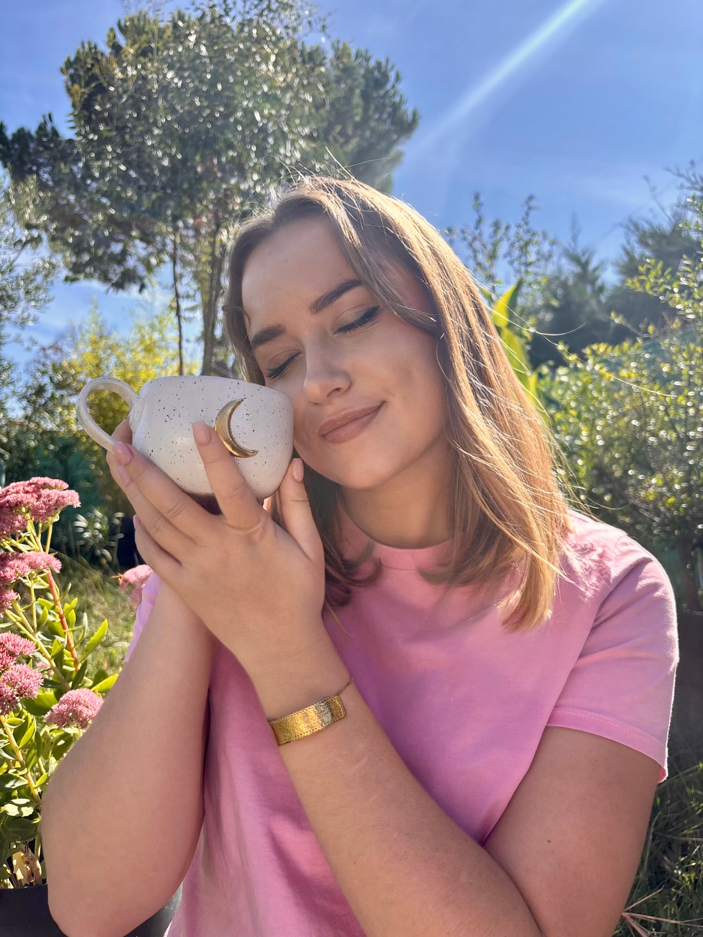 Femme dans un jardin tenant contre sa joue une tasse en grès beige moucheté avec lune dorée