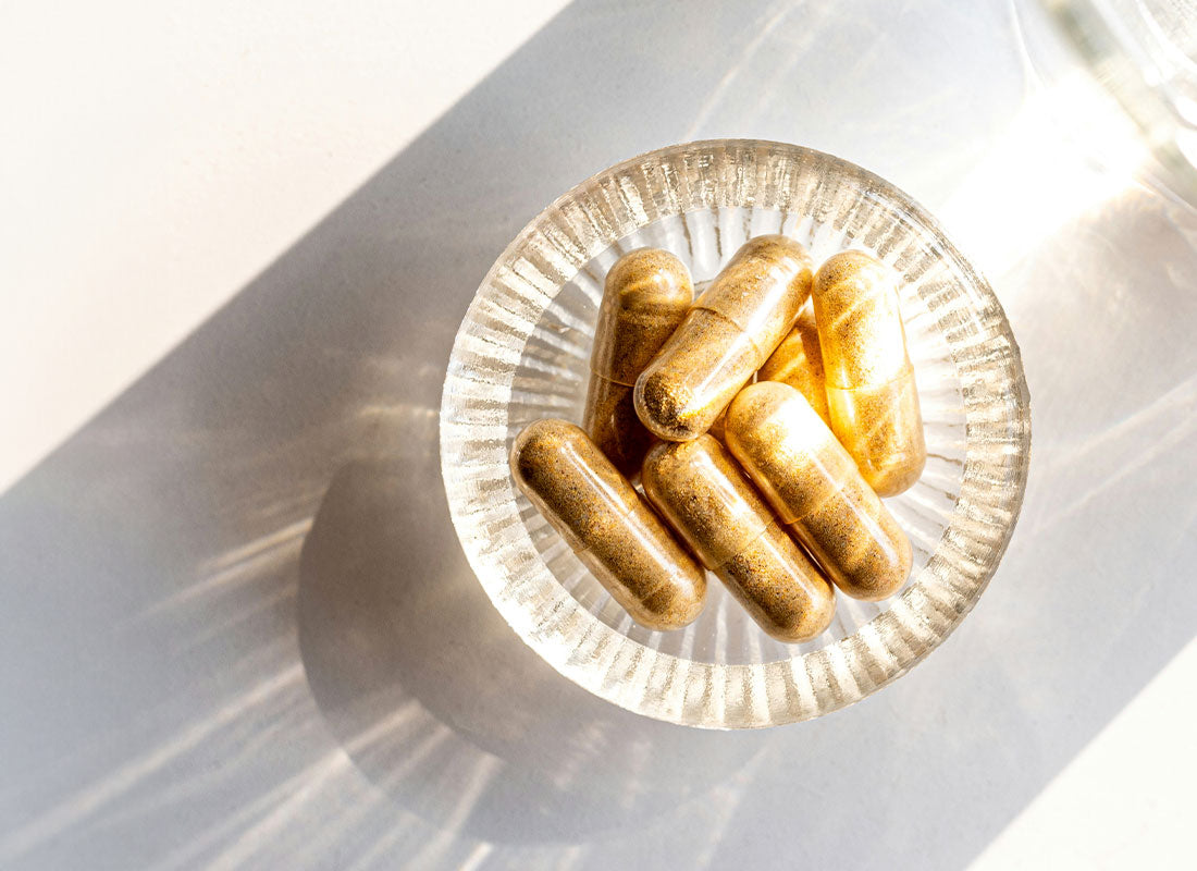 Gélules beiges de compléments alimentaires posées sur une assiette en verre transparent laissant passer les rayons de soleil