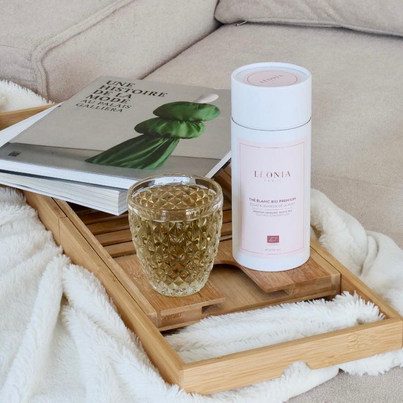 hé blanc bio premium Léonia Paris infusé dans un verre à facettes diamant, posé sur un plateau en bambou avec des livres et un plaid blanc