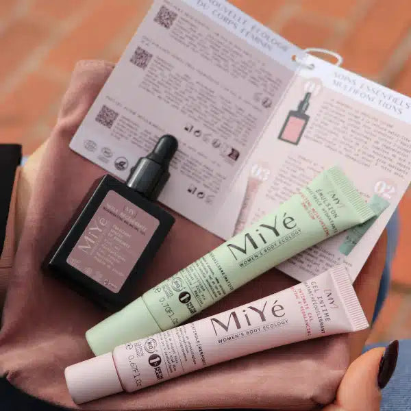 Trio de cosmétiques MiYé, un tube vert de crème visage, un tube rose de gel intime et un flacon avec pipette d'huile sérum visage avec notice explicative dans les mains d'une femme